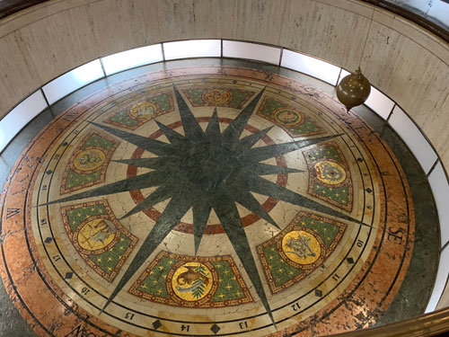 [Elaborately decorated bottom of
the Foucault pendulum, with a compass rose and vague scientific motifs, lit from
a ring of recessed lighting along the bottom.]