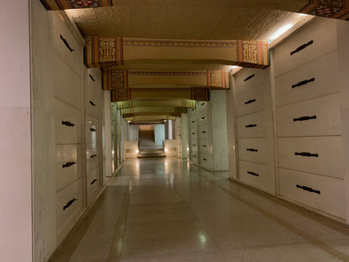 [Hallway of terrazzo floor and
plain stone walls, lined with graves, lit by lighting recessed just under the
ceiling, which is plain with painted crossbeams.]