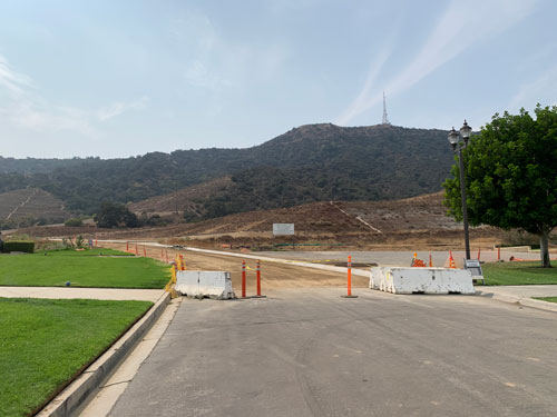 [The paved road and green grass
end abruptly; beyond are mounds of dirt and, past that, the dark chaparral of
the Santa Monica Mountains, with the radio mast visible atop Mount Lee.]